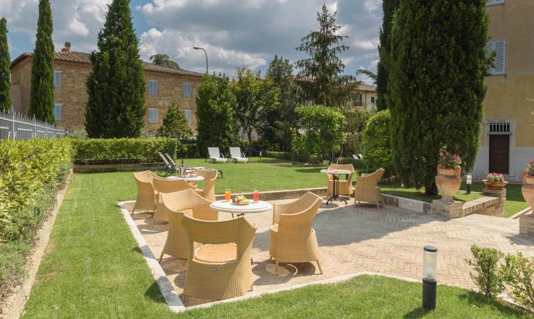 Giardino con sedie in vimini e tavoli, circondato da alberi e cespugli.