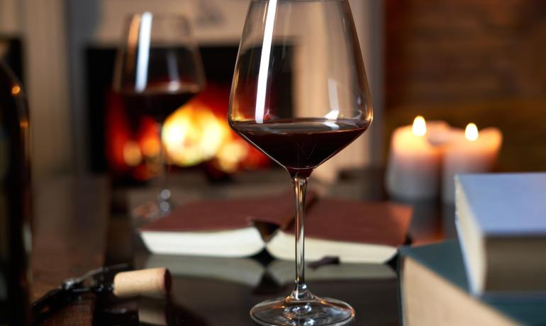 Red wine glass on table with books and lit fireplace.