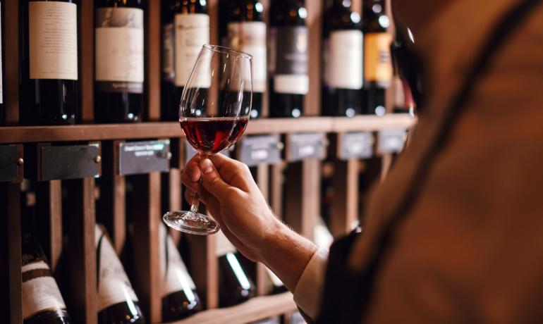 Persona con bicchiere di vino rosso davanti a scaffale di bottiglie.